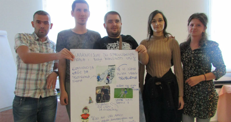 Slika. Na slici je pet mladih osoba, tri mladića i dvije djevojke tokom radionici koji drže flipchart papir sa porukama vezanim za UN Konveciju o pravima osoba sa invaliditetom