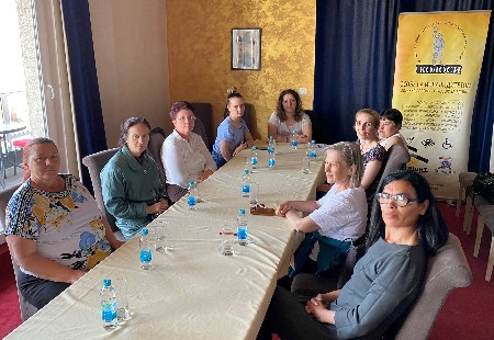Slika nastala tokom sedmog sastanka mentorice sa ženama s invaliditetom u Bijeljini 14. maja, dok sjede za stolom blago okrenute prema osobi koja ih fotografiše, u pozadini je baner Koalicije KOLOSI Bijeljina.