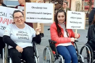 Na slici su dvije članice Foruma žena s invaliditetom Tuzlanskog kantonakoje su u kolicima za osobe s invaliditetom i nose transparente sa natpisima kojima promovišu prava žena s invaliditetom. Slika je nastala tokom osmomartovskih protesta za prava žena u Tuzli.