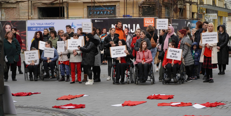 Na slici su članice Foruma žena s invaliditetom Tuzlanskog kantona zajedno sa drugim učesnicama i učesnicima osmomartovskih protesta protiv nasilja nad ženama održanog u Tuzli 8.marta 2024. U rukama nose različite natpise sa porukama koje ukazuju i pozivaju na sprečavanje nasilja nad ženama 